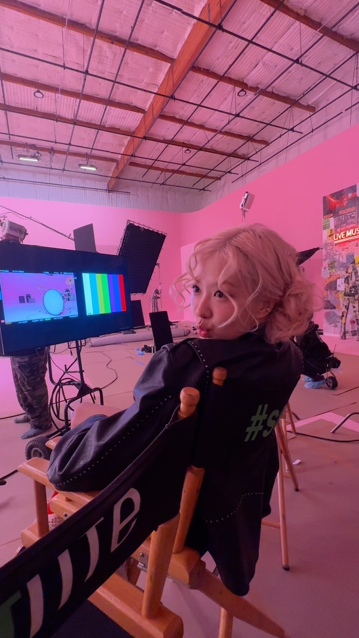 the image shows blackpink's rosé during a music video filming. she's seated in a director's chair, wearing a stylish black jacket with a subtle #s marking. the set is predominantly pink with professional lighting and camera equipment visible.  rosé has blonde hair styled up, and she's wearing makeup.  the background includes monitors displaying video content, suggesting a behindthescenes look at a kpop music video production.