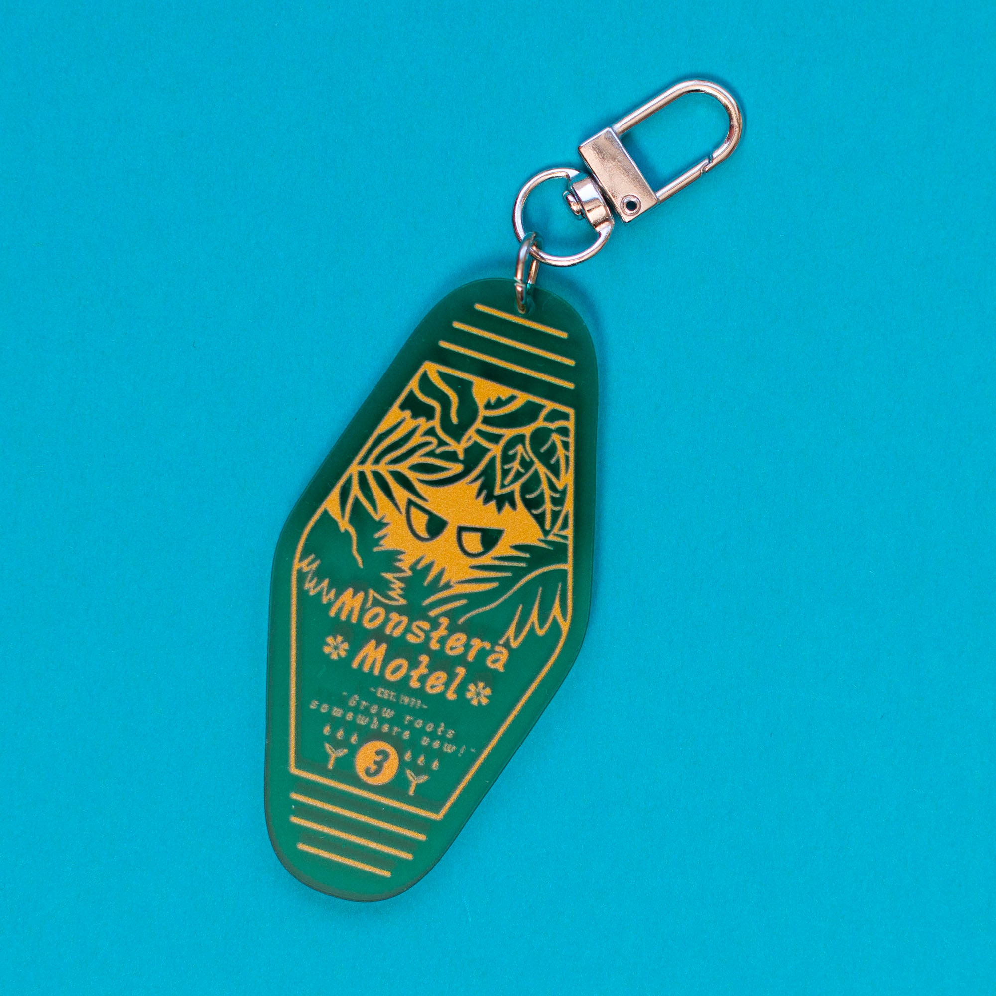 loseup view of a dark green keychain shaped like a vintage motel key tag. the keychain features a stylized illustration of a monstera deliciosa plant with a hidden face in its leaves.  yellow text reads "monstera motel" and includes additional small text. the keychain has a silver split ring for attachment. the background is a bright teal.