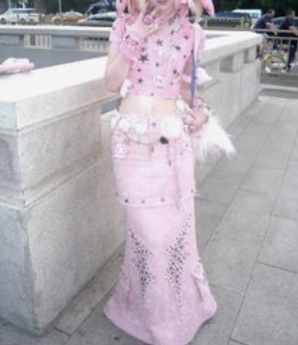 a young woman with pink hair and multiple piercings is posing on a city bridge. she's wearing a pink, y2k inspired outfit that includes a cropped tshirt, a long tiered skirt, and various accessories like belts and necklaces. her style incorporates elements of grunge and kawaii aesthetics. the photo is taken in daylight and features a shallow depth of field, drawing attention to the subject and her outfit.