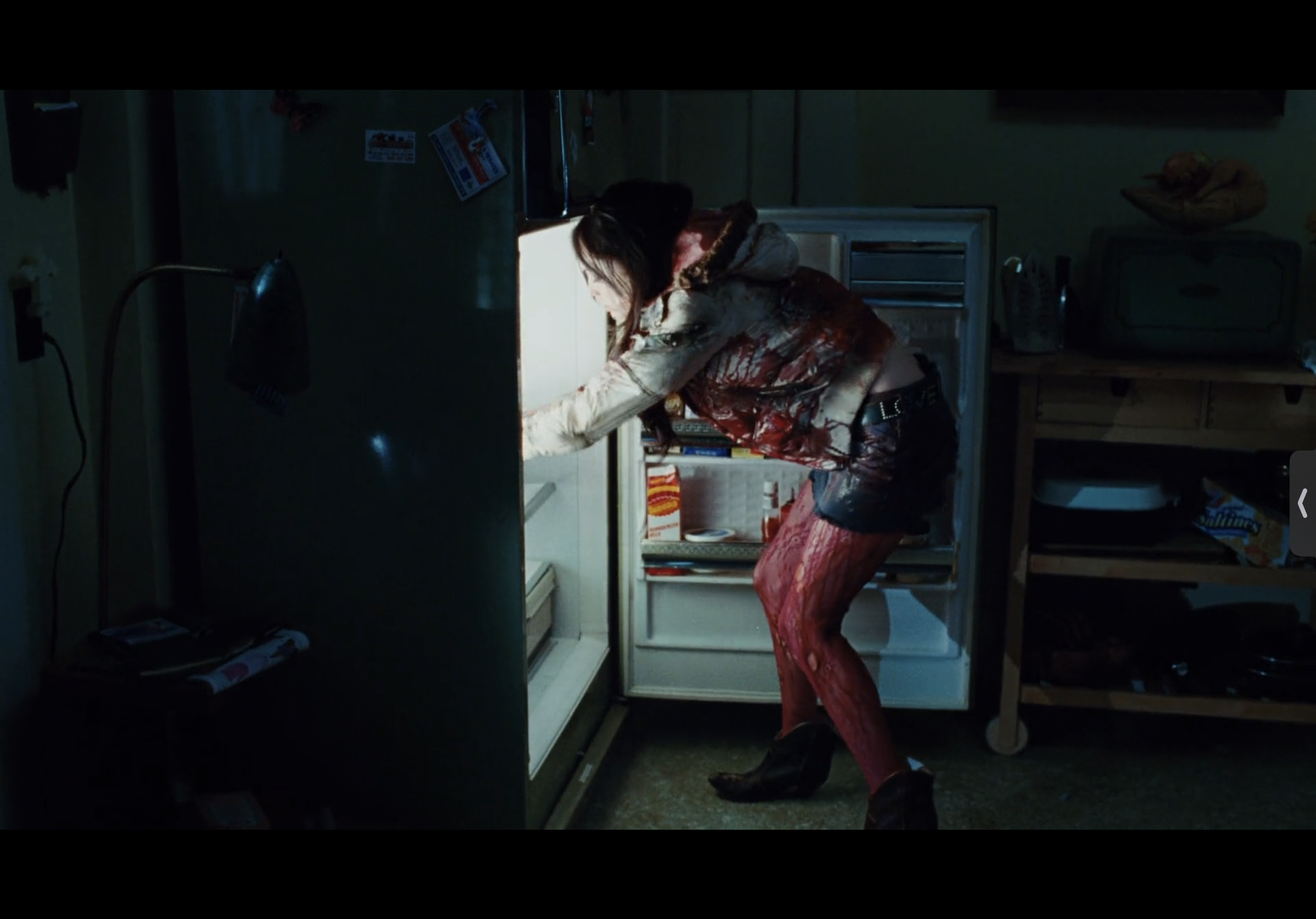 a woman covered in blood stands in a dark kitchen, looking inside an open refrigerator.
