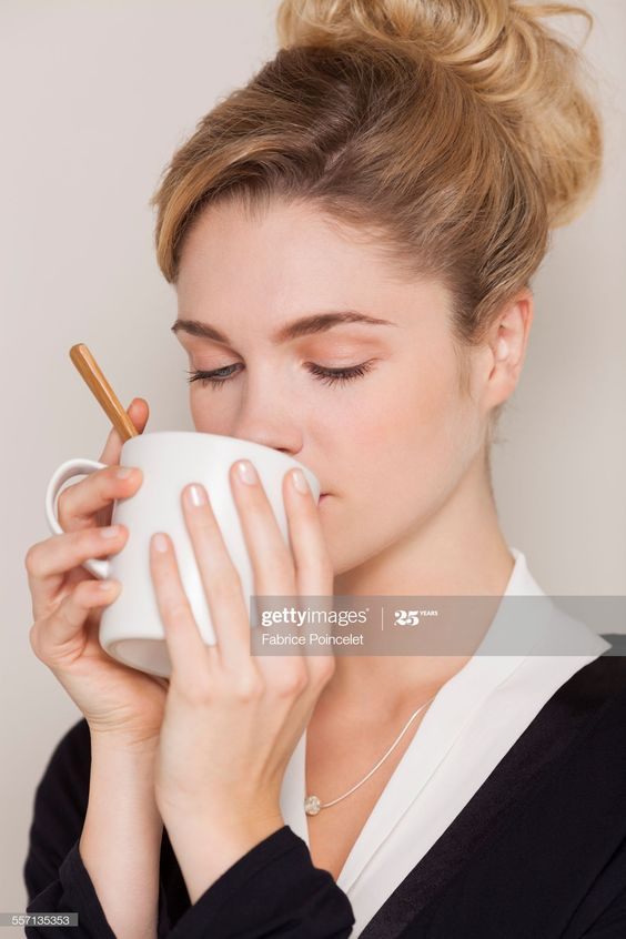 a young woman with blonde hair tied in a bun is shown sipping from a white mug with her eyes closed. she is wearing a black sweater and a delicate silver necklace. the background is a soft beige color. the image suggests a sense of calm and relaxation as she enjoys her warm beverage.