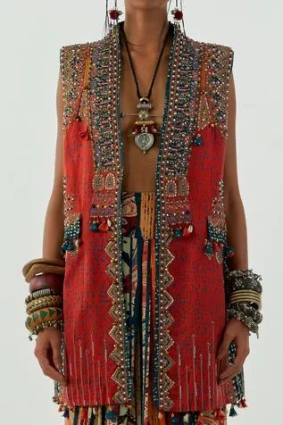 a woman is wearing a red sleeveless vest adorned with intricate beadwork and embellishments, layered over a patterned garment. she is also wearing jewelry, including a necklace and multiple bracelets, enhancing the bohemian, ethnic, and fashionable style of her clothing.