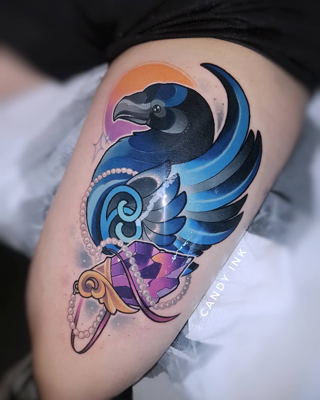a colorful tattoo of a raven on a thigh. the raven has blue and black wings. it is perched on a purple and pink gem that is part of an amulet necklace. the background is an orange circle.  the tattoo was done by candy ink.