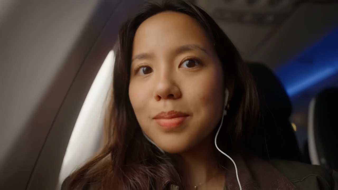 a closeup selfie of a young woman with long dark hair sitting in an airplane seat. she is wearing a green jacket and white headphones, and she is looking directly at the camera with a neutral, content expression. the image has a warm, natural lighting and is taken from a firstperson perspective. the background shows the interior of the airplane cabin, with a glimpse of the window and blue ambient lighting.