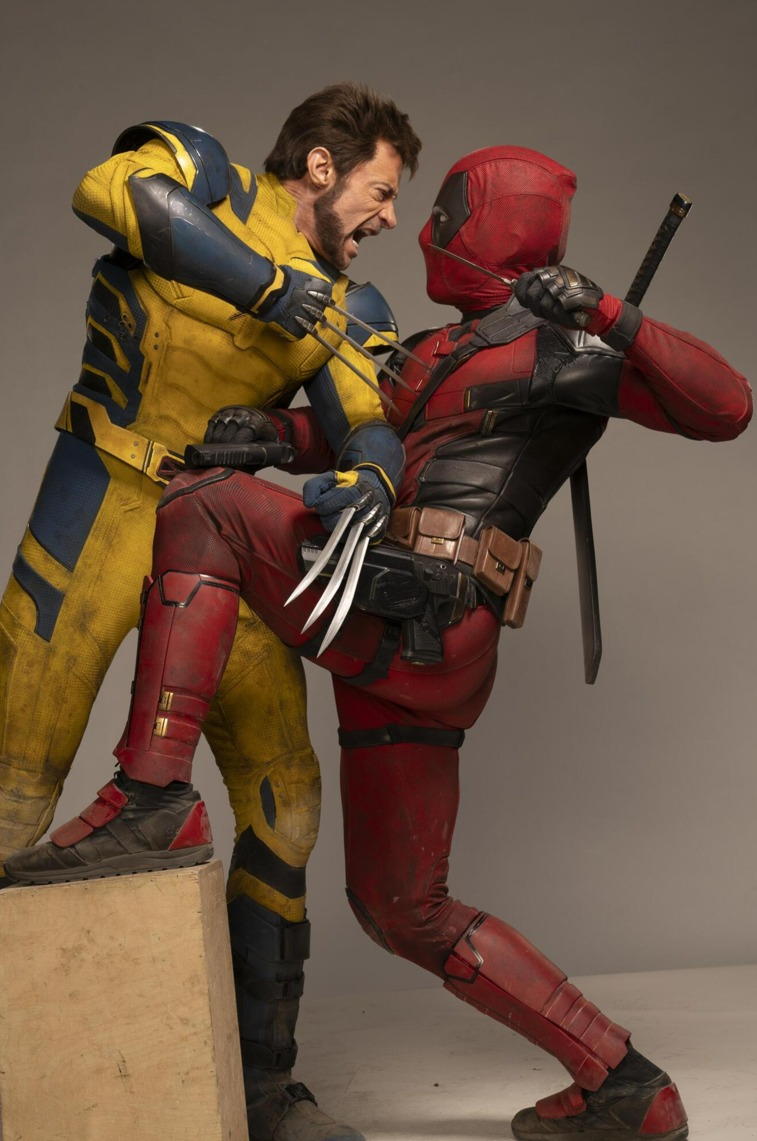 image of hugh jackman as wolverine and ryan reynolds as deadpool seemingly fighting each other. wolverine is wearing a yellow and blue costume with his claws extended. deadpool is wearing his red and black suit, with a sword strapped to his back. they are in a fighting pose, possibly on set.