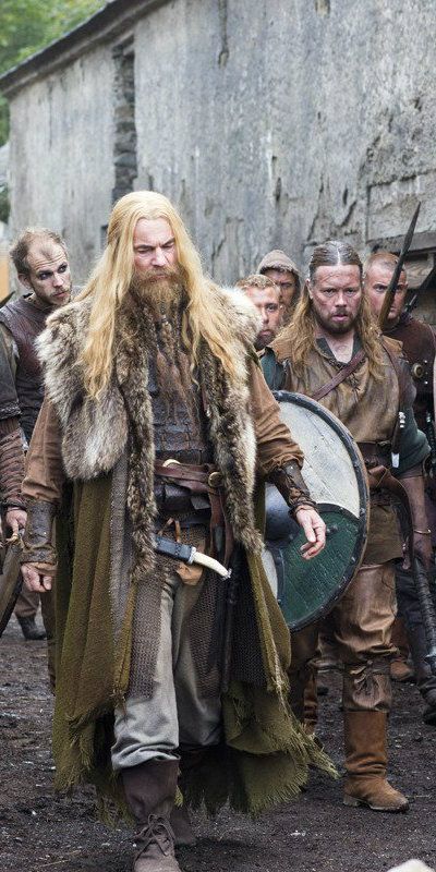 a group of people dressed as vikings are walking outdoors. the person in the foreground has long blonde hair and a beard, wearing a fur cloak and carrying a shield. other vikings are wearing leather armor and carrying spears. they are walking near a building.