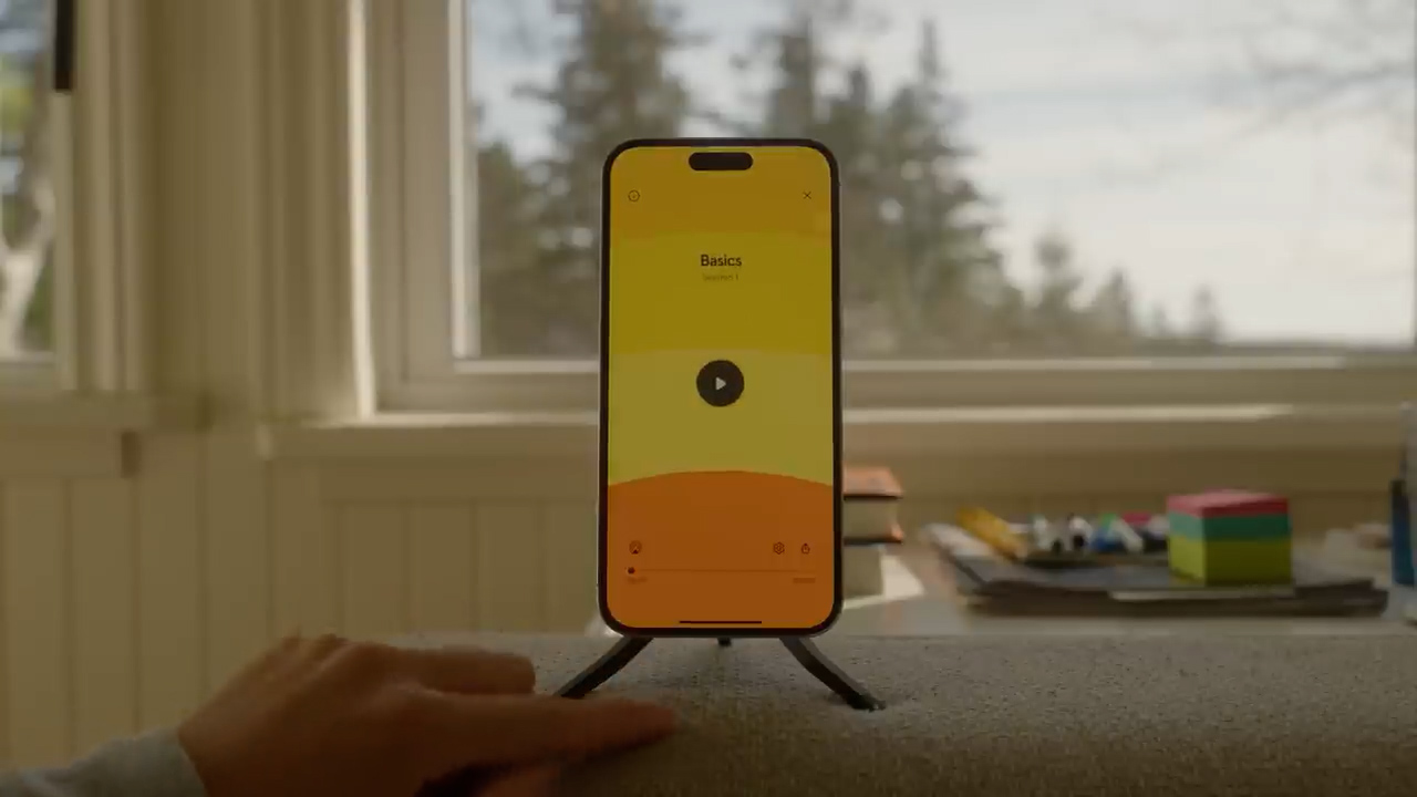 a yellow and white smartphone stands on a table, displaying a music app on its screen. the phone is propped up by a small black stand. a hand reaches out to interact with the touch screen. the background features a brightly lit window showcasing a blurry view of trees and nature, suggesting a serene and comfortable indoor environment. the overall palette is warm and inviting.