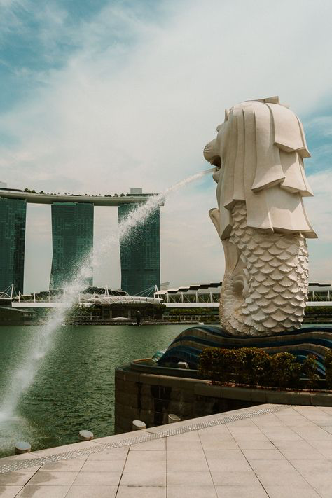 the image shows the merlion statue in singapore, with a fountain of water spraying from its mouth. in the background, the marina bay sands hotel is visible. the scene is set on a sunny day, with the water reflecting the sky. the overall feel is one of travel and tourism, highlighting a popular landmark in singapore.