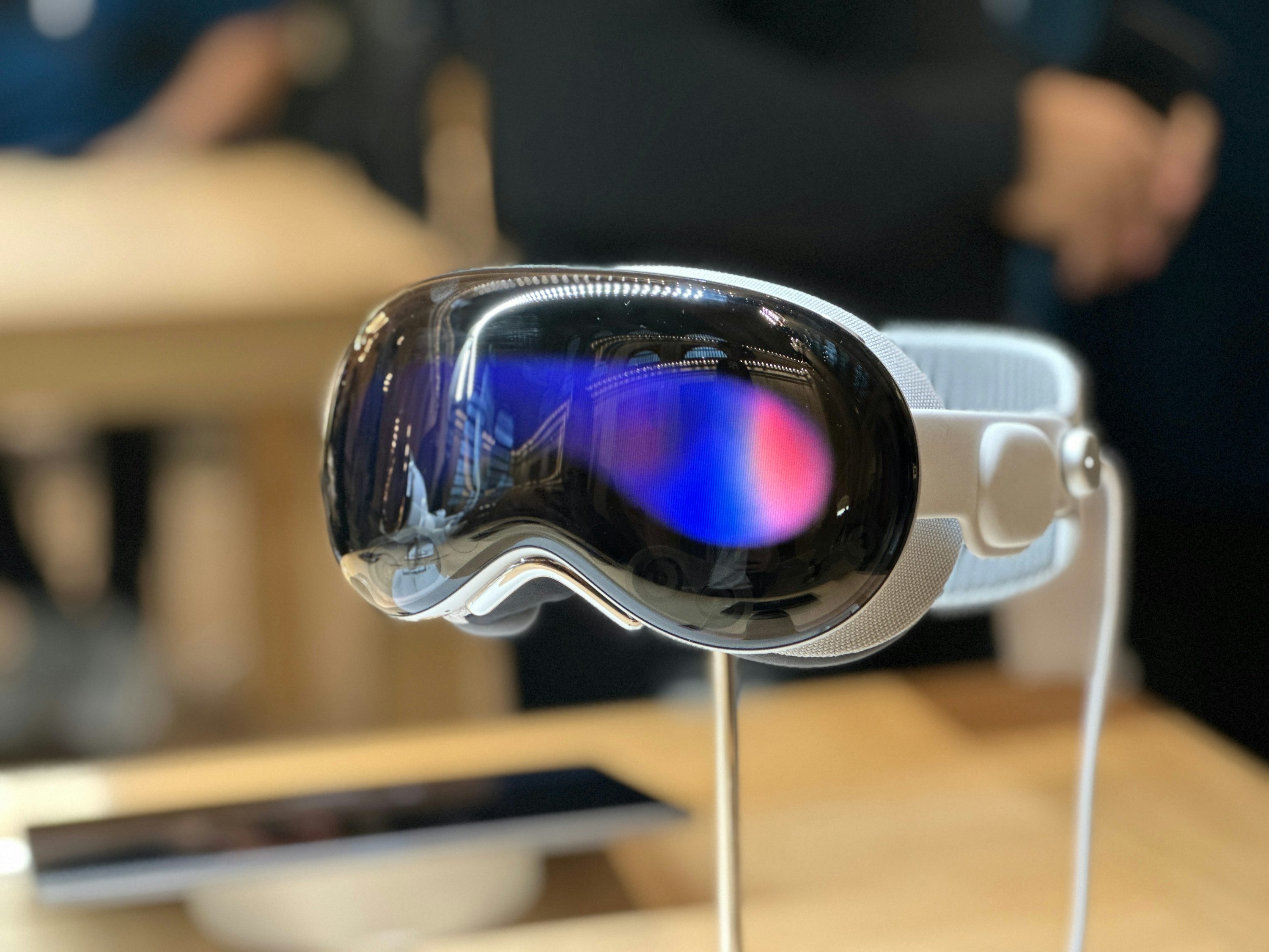 a closeup shot of the apple vision pro headset, showcasing its sleek design, reflective lenses, and overall futuristic appearance. the headset is positioned on a stand in a welllit environment, with reflections visible in the lenses.