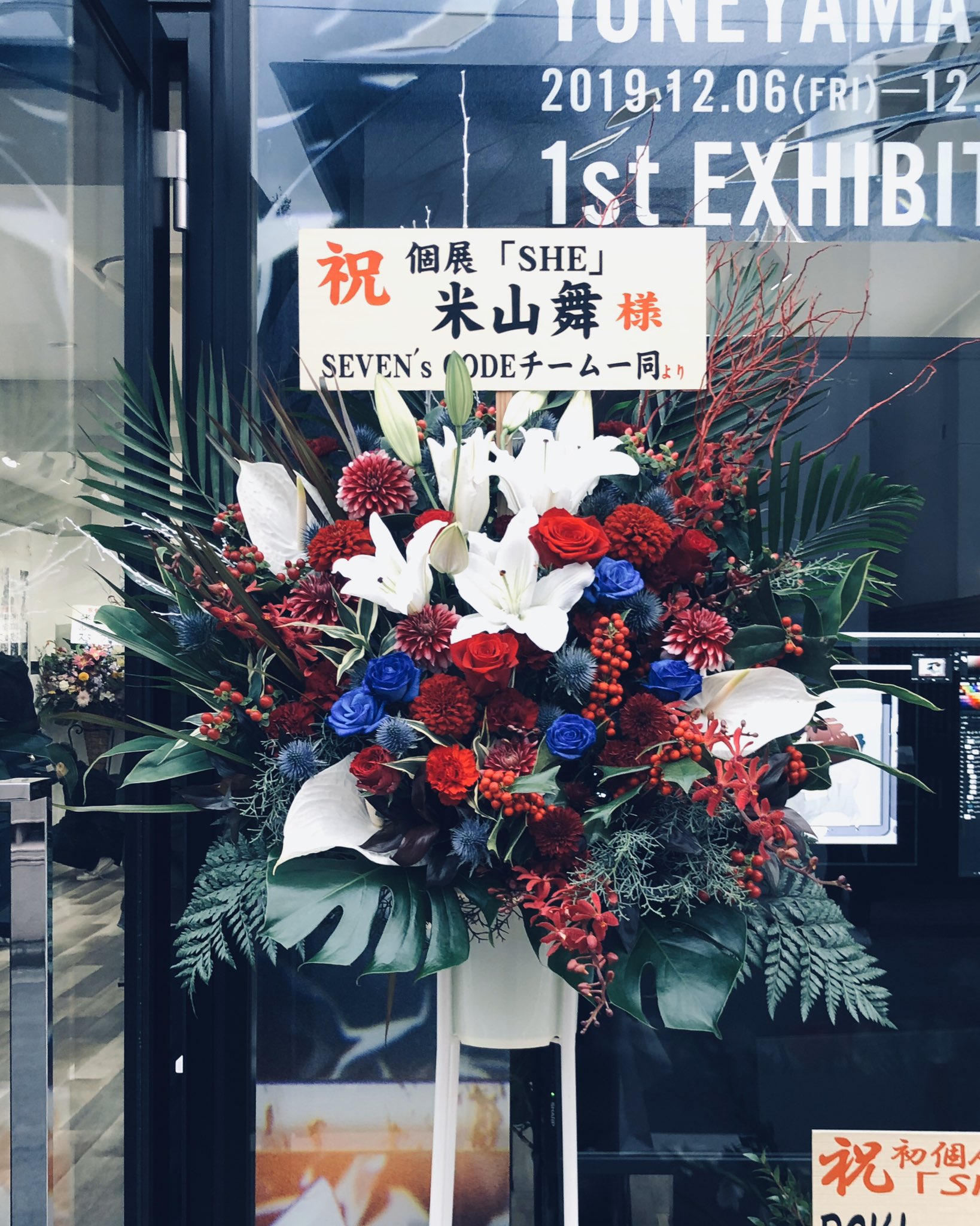 a bouquet of flowers with congratulatory message board in front of a store. the flower arrangement is made of white lilies, red and blue roses and other greenery. the sign is written in japanese with "1st exhibit" in english at the top.