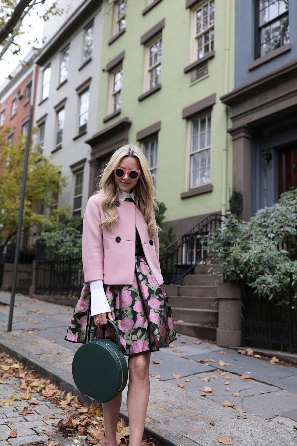 a stylish blonde woman is standing on a city street in front of a row of charming brick townhouses. she's wearing a pretty pink jacket, a dark floral skirt, pink sunglasses, and a dark green round handbag. her outfit is perfect for fall or autumn.  fallen leaves are scattered on the ground around her. the overall aesthetic is sophisticated and fashionable.