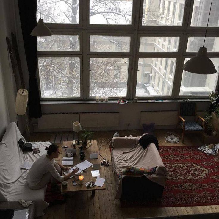 the image shows a highangle view of a loft apartment on a snowy winter day.  large windows dominate one wall, showcasing the snowy cityscape outside. a woman sits at a small wooden table, appearing to work or study, surrounded by books and papers. the apartment is furnished with comfortable couches draped in blankets, a vintage rug, and various plants, creating a cozy and eclectic atmosphere. the overall style is minimalist, yet warm and inviting.  the natural light from the large windows illuminates the space.