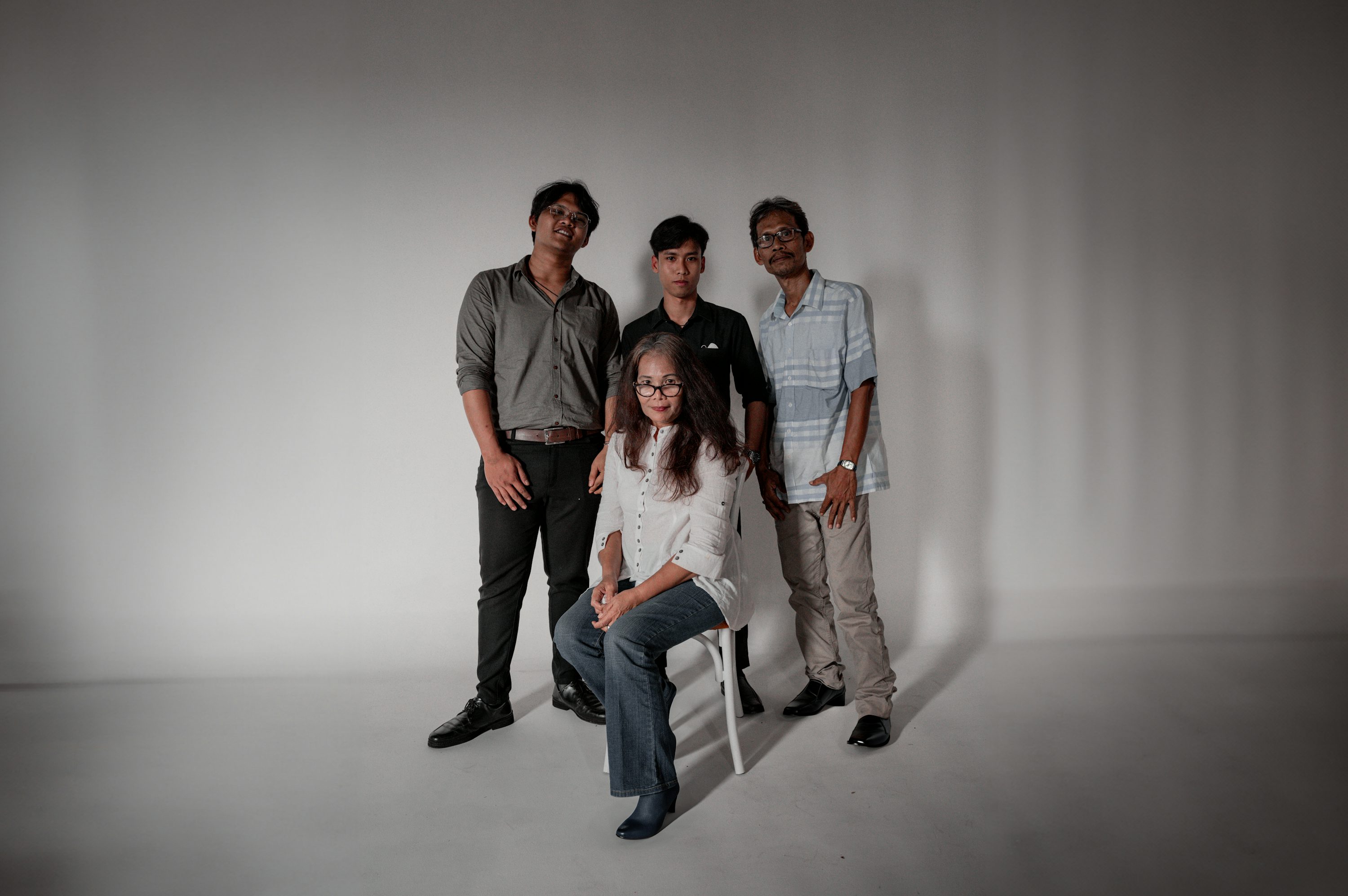 a family portrait of three young men and their mother. the three men are standing behind their mother, who is seated in a chair. the family is smiling at the camera. the background is a plain gray studio backdrop. the lighting is soft and even.