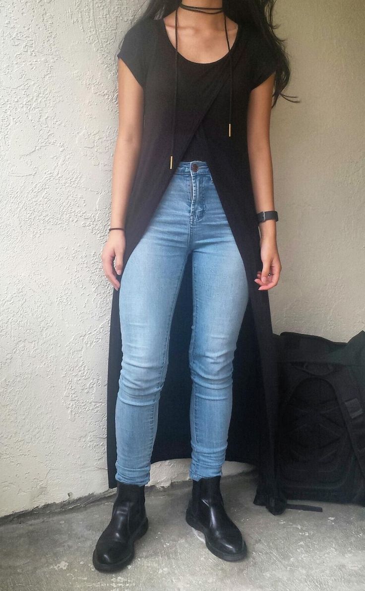 a person is standing in front of a wall wearing a long black dress over light blue skinny jeans and black boots. the person is wearing a black choker. a black bag is behind the person.