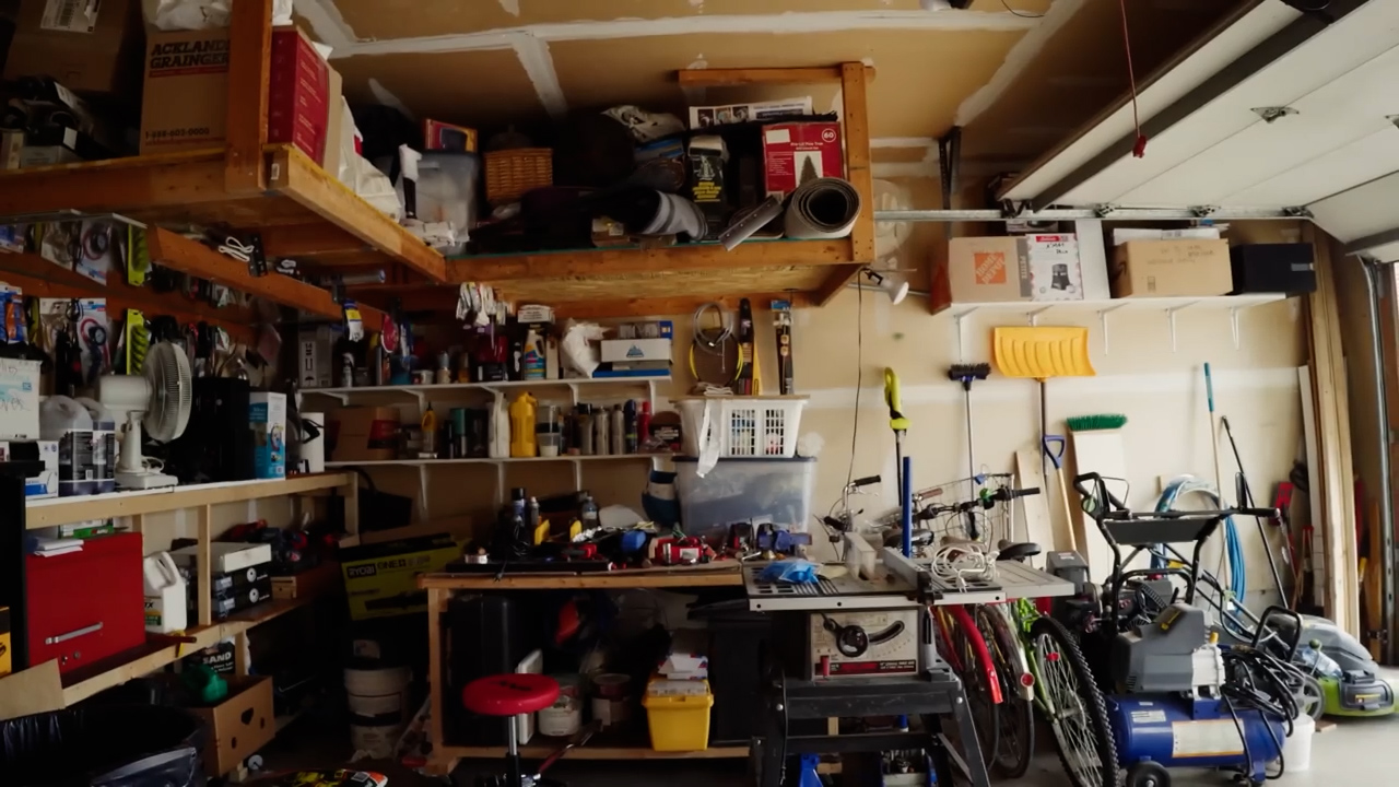 a wideangle view of a cluttered garage interior. wooden shelves line the walls, overflowing with tools, boxes, and various equipment. a workbench sits in the center, also covered in tools and projects. bikes are leaned against the wall, and other miscellaneous items contribute to the overall sense of disorganization. the garage is brightly lit, with natural light streaming in from the open garage door.
