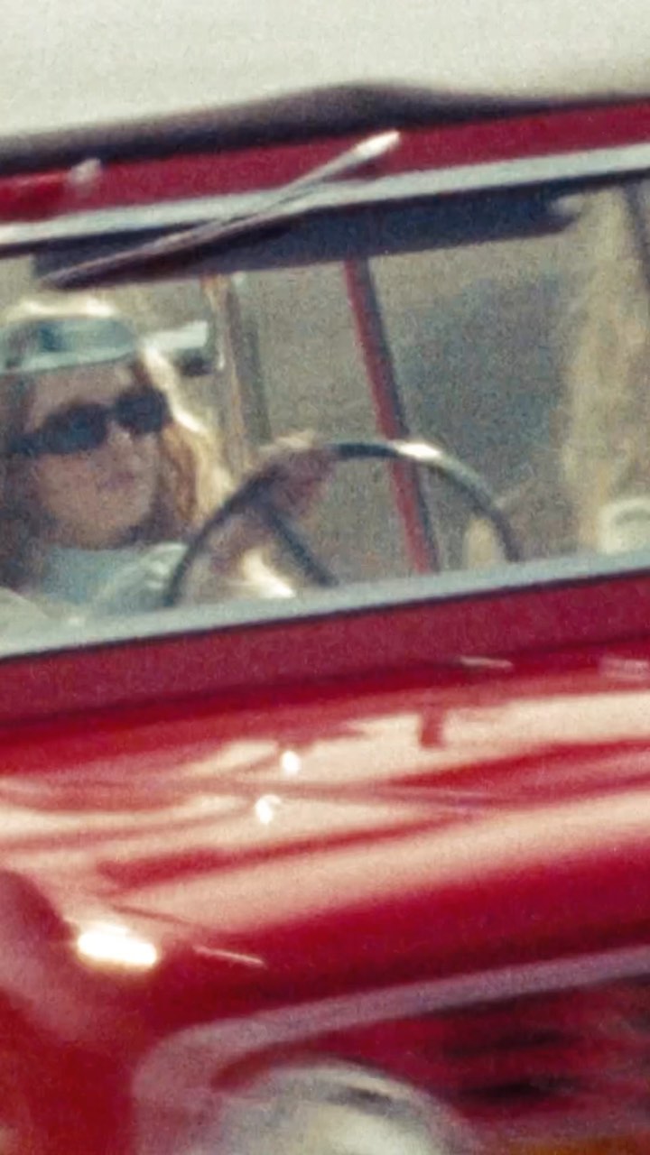 a closeup, grainy photograph appears to be a film still of a woman with sunglasses driving a red vintage ford bronco.  the image is blurry and only shows a partial view of the car and the driver. the car is a classic model, likely from the 1960s or 1970s, and the setting appears to be outdoors during the daytime. the overall feel is retro and nostalgic.