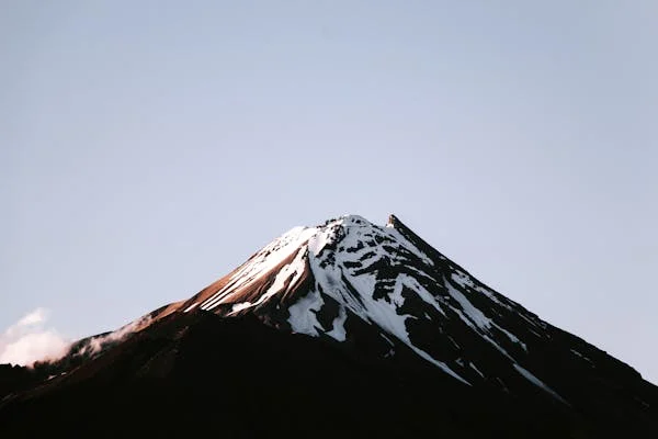 a majestic mountain stands tall against a serene blue sky. its peak is adorned with a pristine blanket of snow, while the lower slopes showcase rugged, earthy tones. the clear daylight casts subtle shadows, highlighting the mountain's grandeur.  a few wispy clouds drift leisurely in the background, adding a touch of ethereal beauty to the scene.