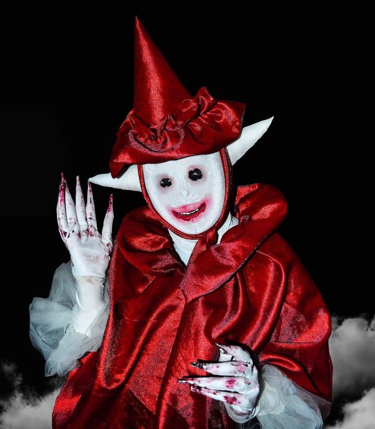 a person wearing a red, pointyhatted elf costume with long, pointy ears and a white mask. the person is smiling, and their nails are long and painted red, with bloodlike streaks on their fingers. the background is dark, with a hint of smoke or fog. the costume is spooky and creepy, and the person's expression is unsettling. this image is likely taken during a halloween celebration, as the costume is a common character during that time.