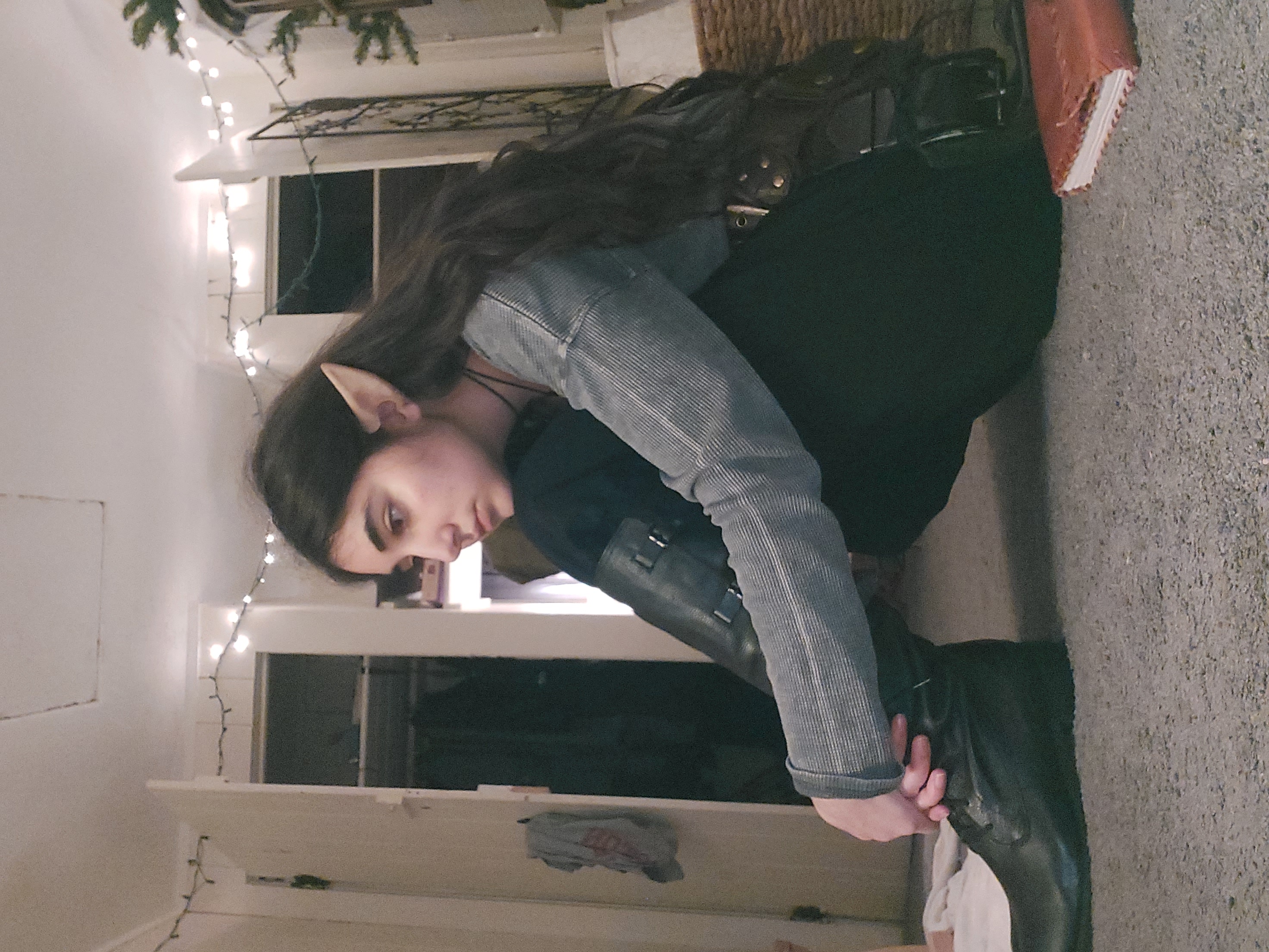 the image shows a teenage girl cosplaying as an elf. she's wearing artificial elf ears and is dressed in darkcolored clothing, including long black boots and a grey longsleeved shirt.  her long brown hair cascades down her back. she is sitting on the floor of what appears to be her bedroom, which is dimly lit with string lights. she looks pensive or thoughtful, her gaze directed downward. a journal or book lies on the floor near her. the overall mood of the photo is moody and contemplative. the background shows a partially visible closet and some other bedroom furniture.