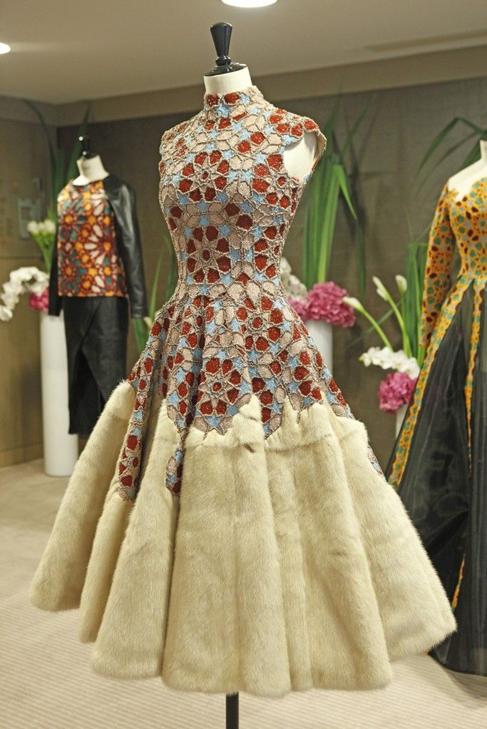a mannequin wears a sleeveless dress with a high neck. the top of the dress features a geometric floral pattern in red, blue, and beige colors. the skirt is made of beige fur and has a flared shape. other mannequins and clothing are visible in the background, along with floral arrangements.