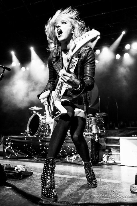 a black and white image of a female rocker performing on stage. she is playing an electric guitar and singing into a microphone. she is wearing a leather jacket, black boots, and black pants. the image is taken from a slightly low angle and captures the energy of the performance.