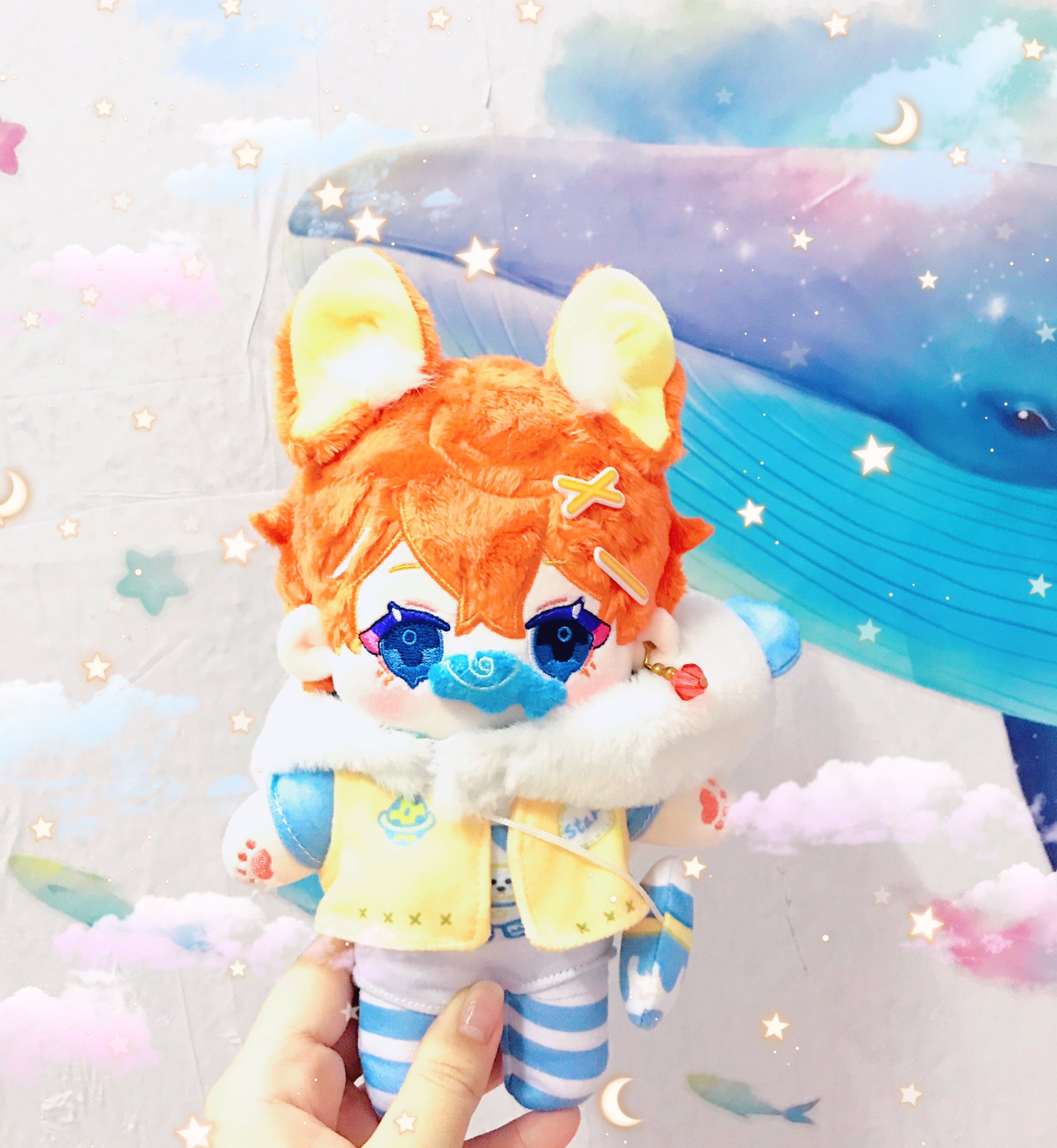 a plush toy of an anime character with orange hair, blue eyes, and a cute outfit. it is wearing a yellow vest over a white shirt, blue and white striped pants, and a white fur collar. the toy is being held in someone's hand against a background of clouds and stars.