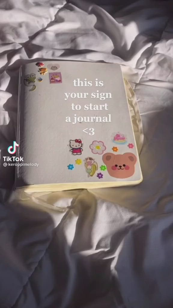 a closeup shot of an open journal laying flat on a soft, white, fluffy blanket. the journal has a white cover and is decorated with cute pastel stickers. the text on the page reads "this is your sign to start a journal <3". the photo is slightly blurry because of the blanket.