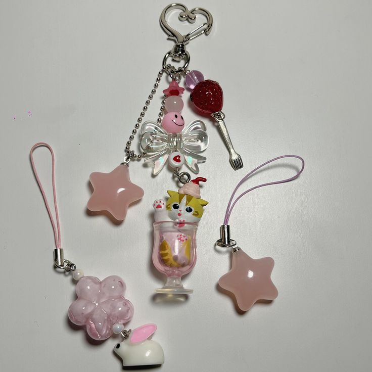 the image shows a keychain with several charms hanging from it. the keychain has a heartshaped clip. the charms include pink stars, a beaded pink flower with a white bunny, a cat in an ice cream cup, a pink bow with a smiley face, a strawberry on a fork, and pink and purple beads.