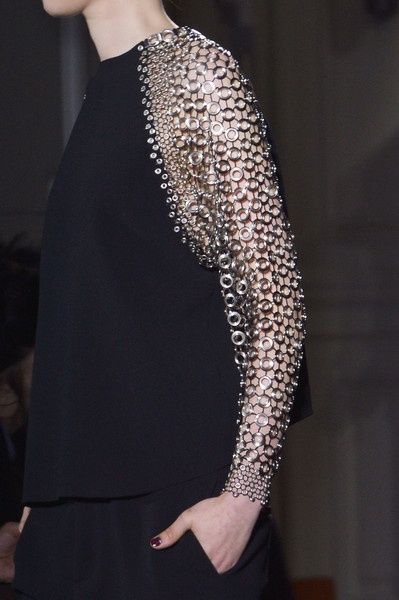 a woman wearing a black blouse with a sleeve made of linked metal circles. the garment is an example of high fashion, possibly designer clothing for a runway show.