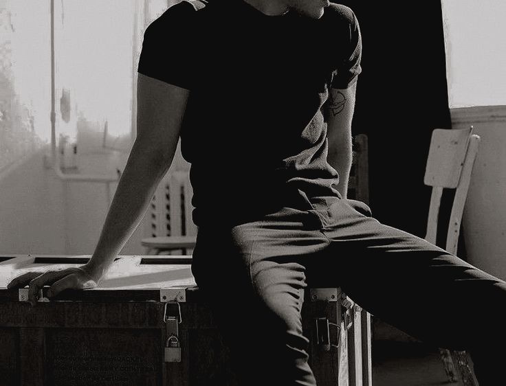a man in a black shirt and pants is sitting on a crate in an abandoned building. the image is in black and white, with a moody and gritty feel. the man's silhouette is against the light coming in from the window. the chair in the background adds to the sense of emptiness and solitude. the image evokes a feeling of loneliness and isolation.
