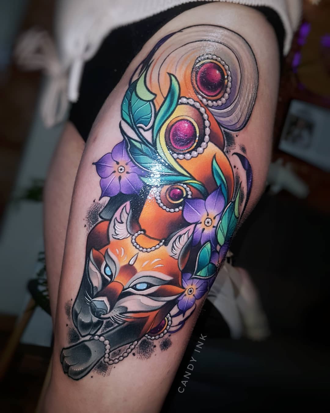 a closeup view of a colorful neotraditional tattoo on someone's upper thigh. the tattoo depicts a stylized fox in orange and black curled up with purple flowers near the top. the fox has blue eyes and is adorned with pearls and colorful gems.  the tattoo artist's signature, "candy ink" is visible in black near the bottom right.