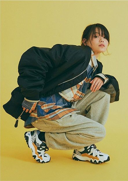 a young woman poses against a vibrant yellow background. she is styled in a casual, trendy outfit consisting of an oversized black bomber jacket, a blue and orange plaid shirt, grey sweatpants, and black and white chunky sneakers.  the low camera angle emphasizes her confident stance and highlights the details of her outfit, particularly the footwear. the studio lighting casts subtle shadows, enhancing the overall visual appeal of the image.
