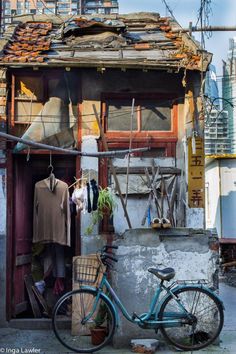 a weathered, dilapidated house in an urban setting. the exterior is a mix of brick and wood, showing signs of age and disrepair. a bicycle leans against a low wall in the foreground. laundry hangs on a line strung across the front of the house, and a few potted plants add a touch of greenery. the scene captures a sense of everyday life in a densely populated urban environment.
