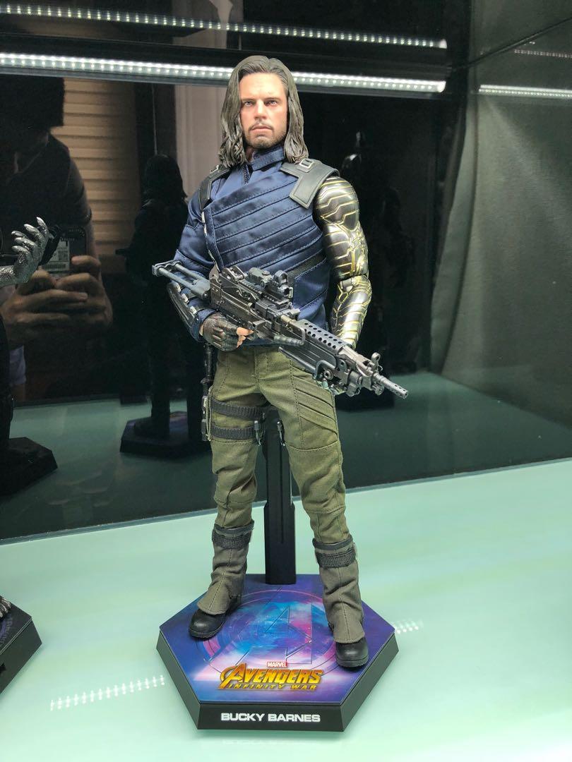 a closeup shot of a highly detailed 1/6 scale action figure of bucky barnes (winter soldier) from marvel's avengers. the figure is accurately depicted in his endgame attire, complete with his signature metal arm and long hair. he's holding a machine gun and is displayed on a hexagonal stand with the avengers logo and "bucky barnes" inscribed on it. the background is blurred, suggesting a display case or shelf.  the lighting accentuates the figure's details and color.