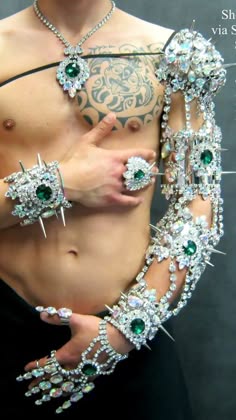a man is wearing a sparkling silver  arm bracelet with green gems and a diamond necklace with a green gem pendant. he has tattoos on his body.