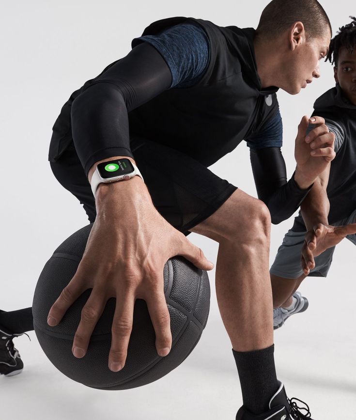 the image showcases two young men engaged in a dynamic workout. the focus is on the man in the foreground, who is wearing a black athletic vest and compression sleeves, and an apple watch series 8 on his wrist. he is holding a medicine ball, and his pose suggests a moment of intense physical activity.  the second man is slightly blurred, positioned in the background and also dressed in athletic gear. both are clearly focused on their workout, highlighting the aspects of fitness, health, and technology showcased by the apple watch. the scene is set against a stark white backdrop for clean visual impact.