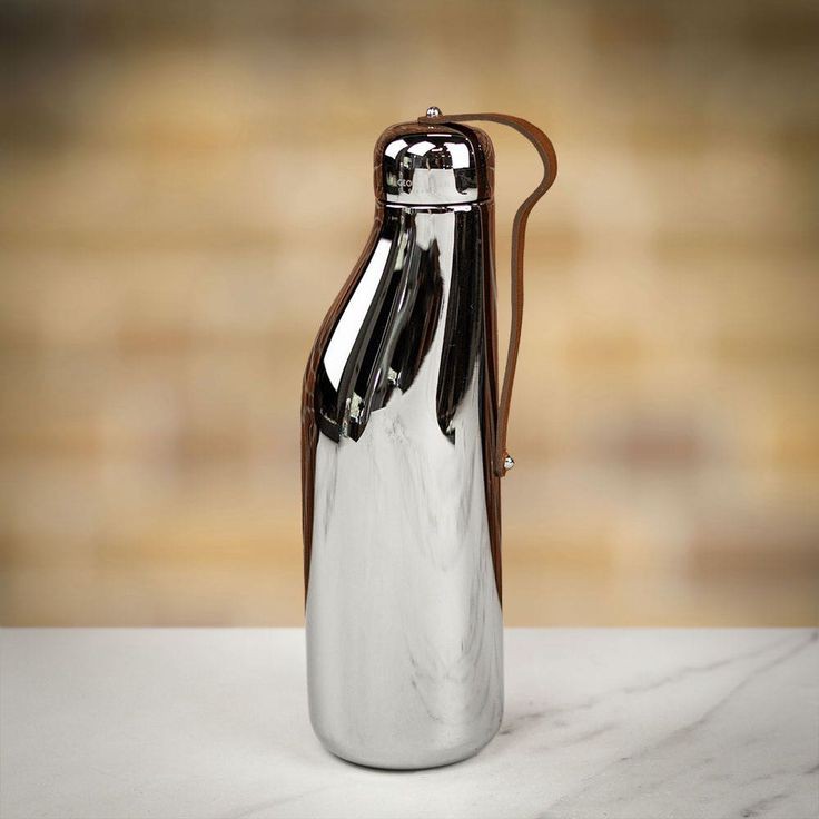 a shiny, silver metal water bottle with a brown leather strap stands on a white marble surface. the bottle has a unique, curved shape and a mirrored finish reflecting the surroundings. the background is blurred with a warm, neutral tone.
