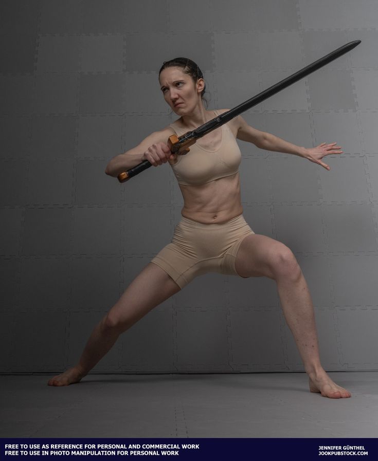 the image shows a muscular woman in a dynamic fighting pose. she's holding a long, dark sword with both hands, her body angled, suggesting movement and attack. she's wearing a light beige sports bra and shorts. the background is a simple gray textured wall.  the overall feel is one of strength, power, and action, making it suitable as a reference image for artists, animators, and others in need of dynamic female poses.