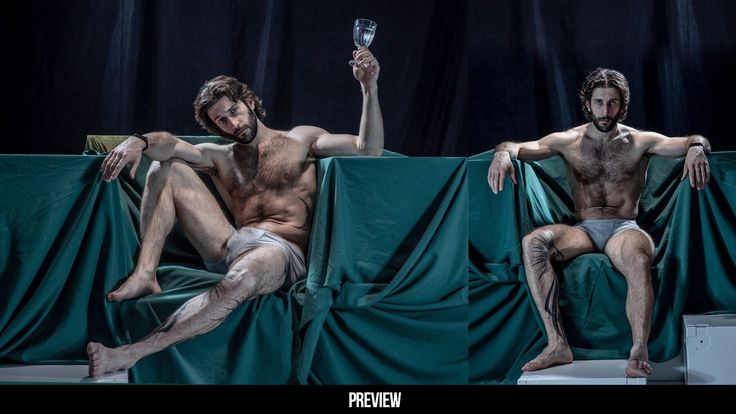 two shirtless men sit on the same teal fabric chair in a studio setting. they are posed in a symmetric fashion that shows the same pose, one is holding a glass of wine.