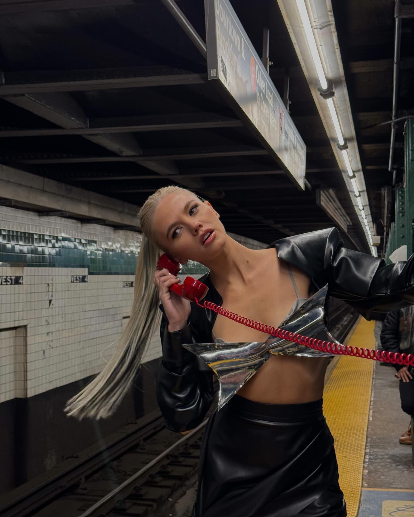 a woman stands on a subway platform talking on a red corded telephone. she is wearing a black jacket and skirt, and a shiny silver top that looks like a bow. the subway platform is yellow and there are tracks visible. the wall behind her is tiled.