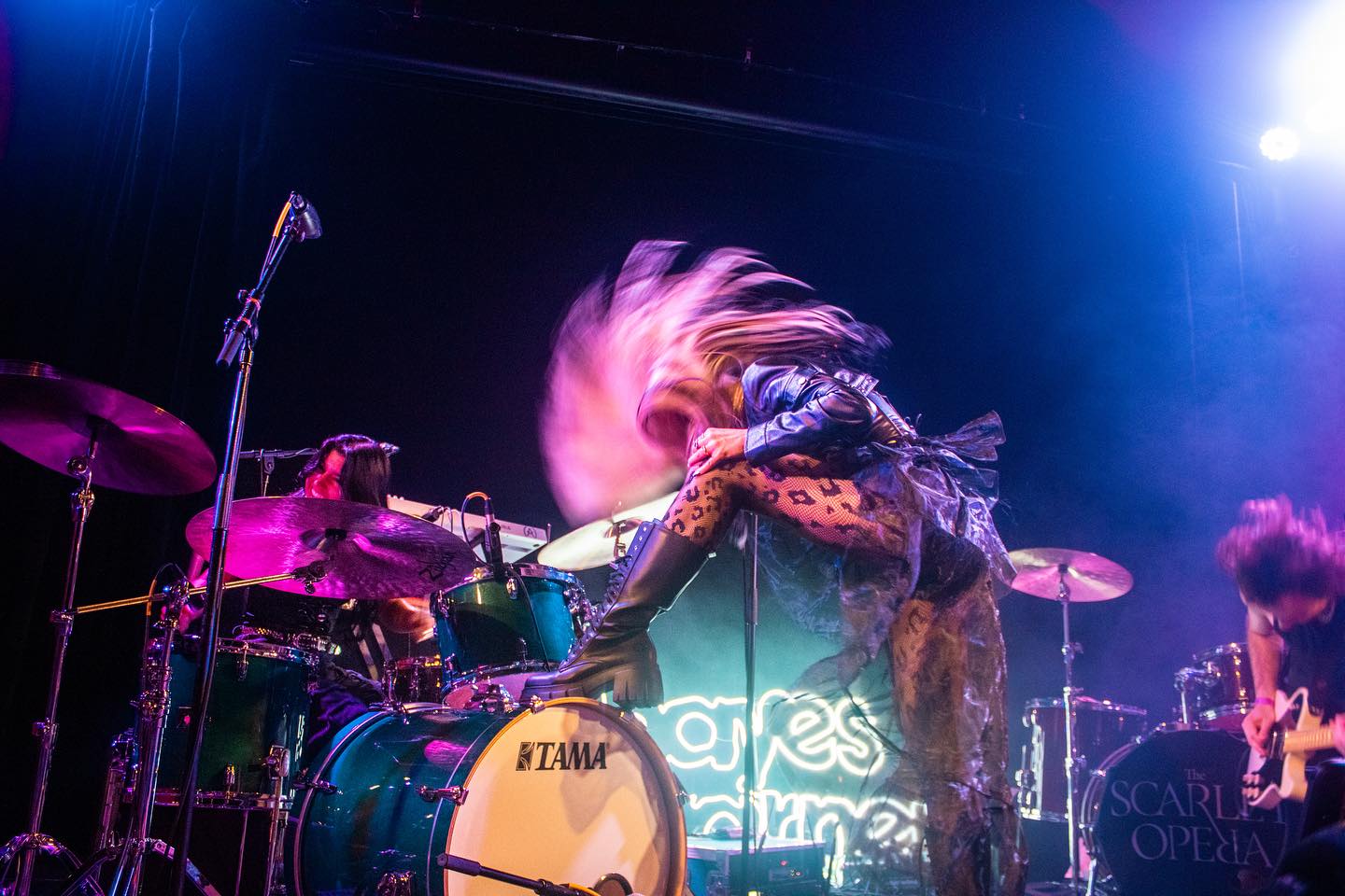 this is a concert shot of the music group the scarlet opera playing live on stage. the drummer is headbanging and kicking the drum set. there is also a guitarist on stage. there is a singer in the background behind the drummer.