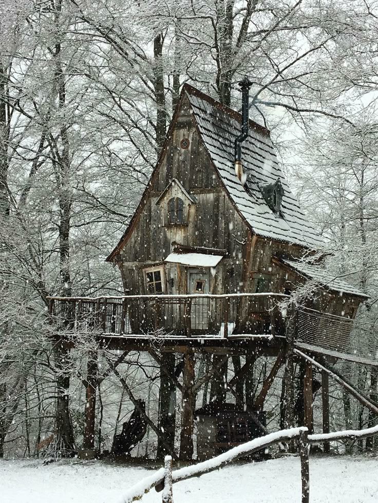a whimsical wooden treehouse stands in a snowy forest. the rustic cabin features an aframe design with a tiled roof, a chimney, and is elevated on wooden stilts. snow is falling heavily, blanketing the ground and the surrounding trees. the structure has a balcony and a small window, and a wooden fence is visible in the foreground.