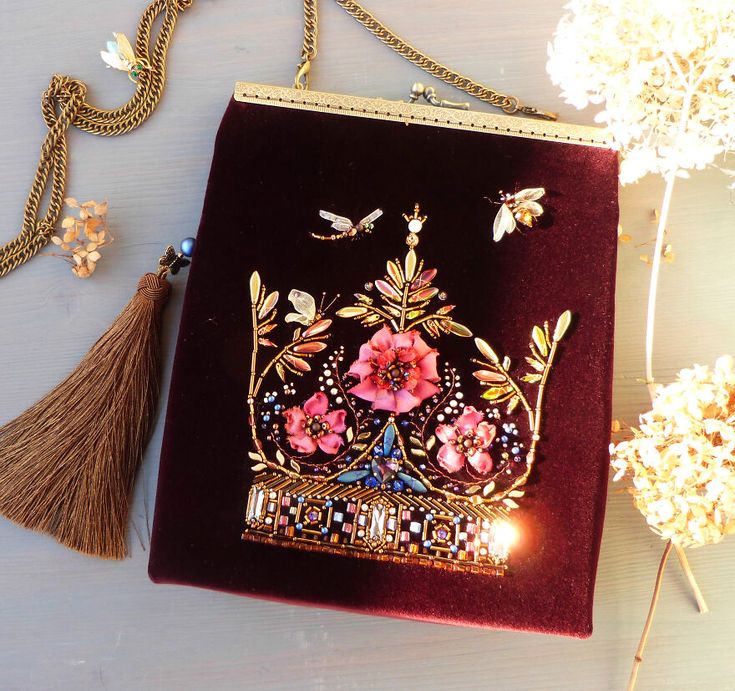 this image shows a maroon velvet clutch bag with a gold chain strap and tassel. the bag is adorned with a beaded embroidery that features a floral motif, a crown, and two dragonflies. it has a vintage style and is a handmade, elegant fashion accessory. the bag is sitting on a light grey surface, and there are some dried flowers in the background.