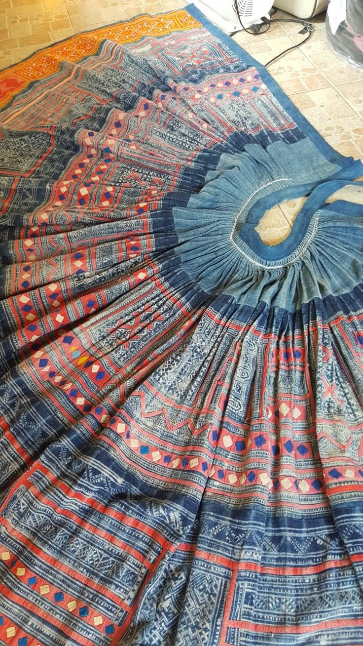 the image shows a pleated skirt made from a colorful textile. the fabric has a complex pattern of blue, red, and white, including stripes, geometric shapes, and embroidery. the skirt has a pleated waist and is lying on a tile floor. the overall impression is of a traditional, handcrafted garment.