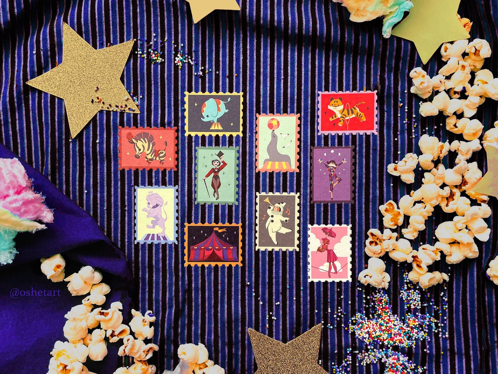 a flatlay image showcasing a collection of whimsical circusthemed stamps arranged on a dark blue striped fabric.  the stamps depict various circus acts and animals, including a seal balancing a ball, an elephant, a zebra, a tiger, a lion, a ringmaster, an acrobat, and a clown.  scattered around the stamps are popcorn, gold glitter stars, and cotton candy, creating a festive and playful atmosphere. the overall aesthetic is vintage and retro, with a slightly handdrawn illustration style. the image is brightly lit and wellcomposed, making it suitable for social media or marketing purposes.  a watermark "@oshetart" is visible in the bottomleft corner.