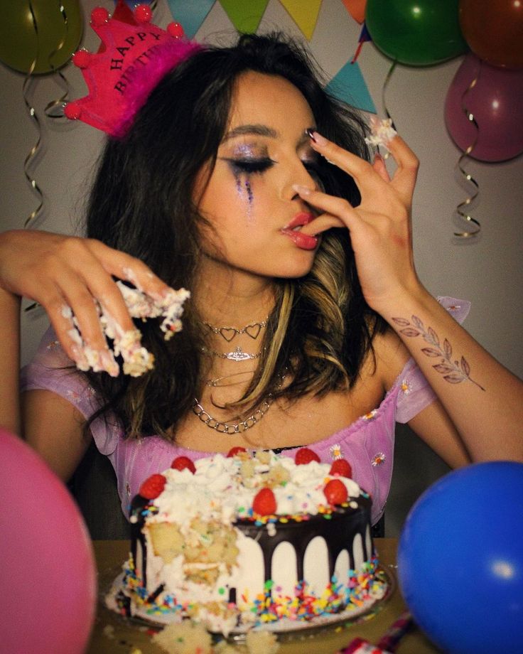 the image shows a young woman with dark hair wearing a pink "happy birthday" crown, sitting at a table and messily eating a multilayered birthday cake decorated with strawberries, sprinkles, and chocolate ganache. she has glitter makeup on her eyes and is wearing several necklaces. there are pink and blue balloons in the background, typical of a birthday party scene. the overall mood is playful and celebratory, with the focus being on the happy, but slightly chaotic, enjoyment of the cake.  a leaf tattoo is visible on her wrist.
