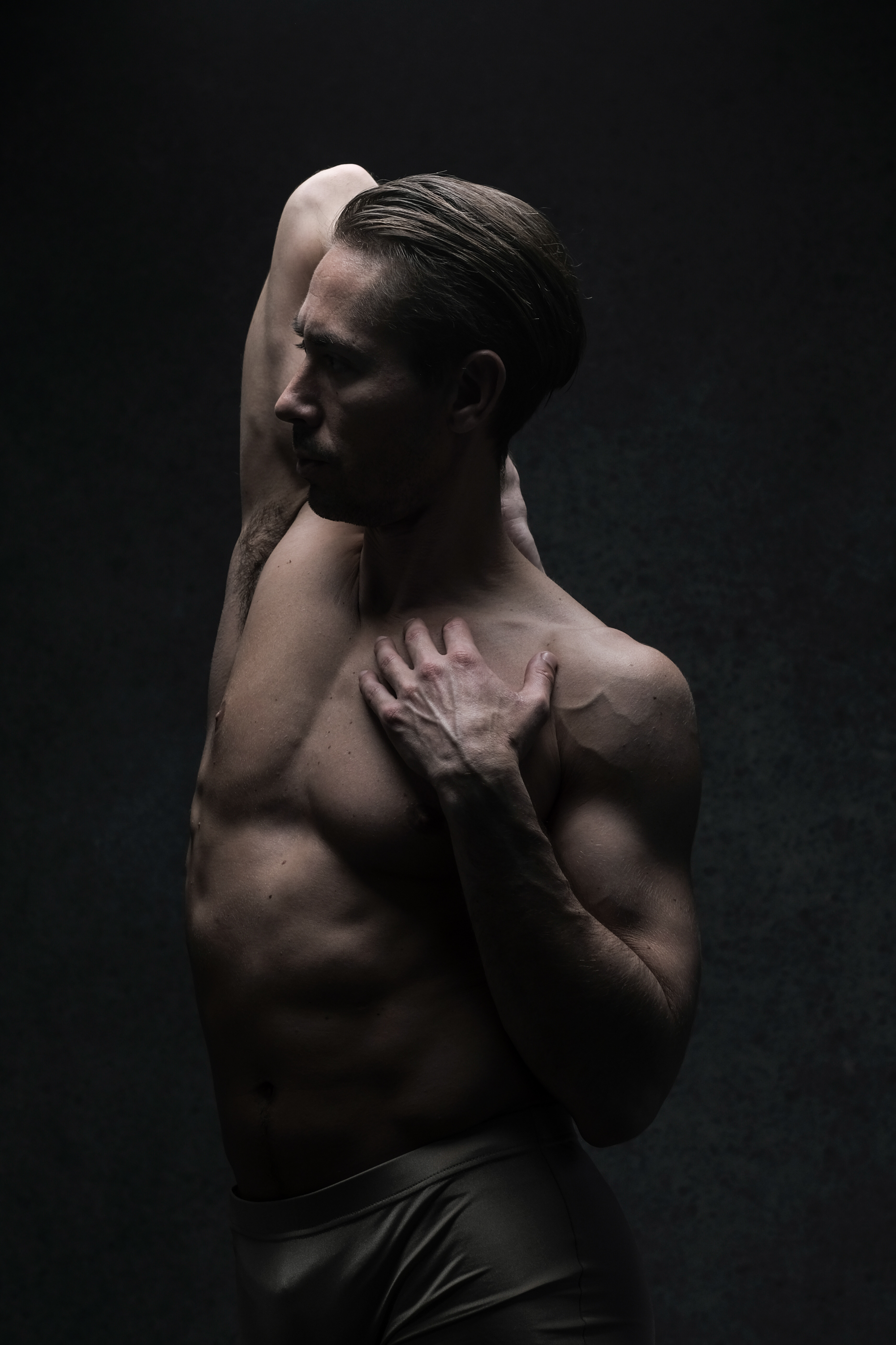 a shirtless male dancer in gray underwear is shown in profile against a dark gray background. his right arm is raised above his head, and his left hand rests on his chest. the studio lighting highlights the contours of his muscular body.