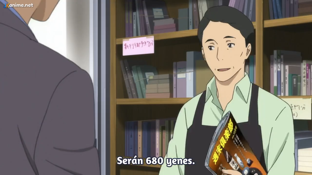 this is a scene inside a bookstore. a man wearing an apron, likely a bookstore worker, is showing a magazine to a customer. the customer's back is to the camera. the bookstore worker is smiling.  there are bookshelves filled with books in the background.  this scene is lit with indoor lighting.