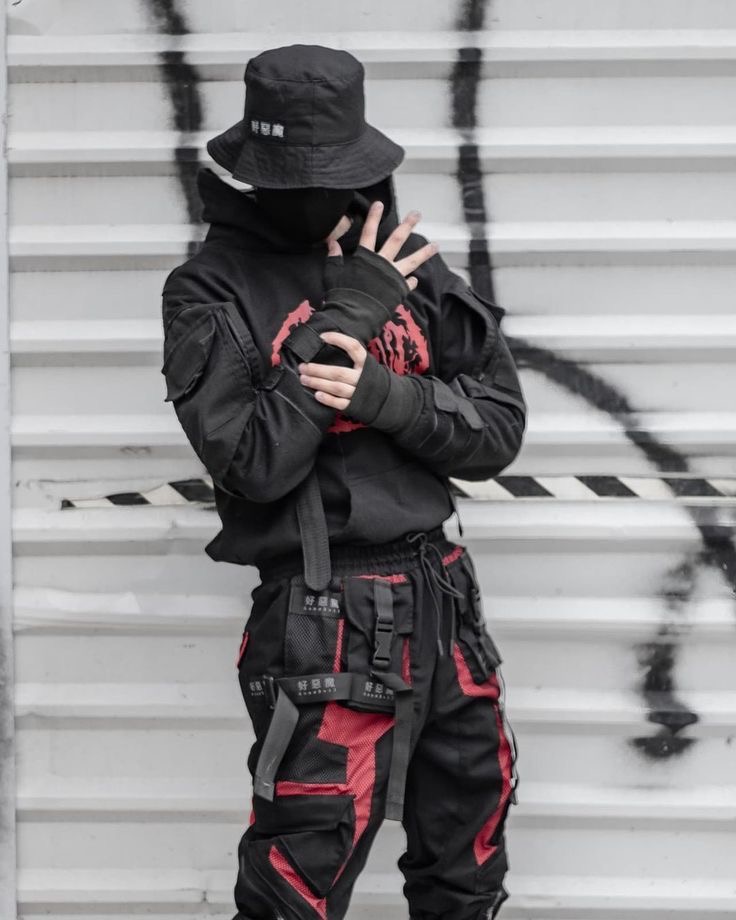 a man is standing in front of a white metal fence, wearing a black hoodie, black cargo pants with red accents, and a black bucket hat with black mask. he's posing with his hands near his face and looking away from the camera. he's wearing several accessories, including a silver chain and a black belt. he's giving off a cool and edgy vibe with his fashion sense and pose.