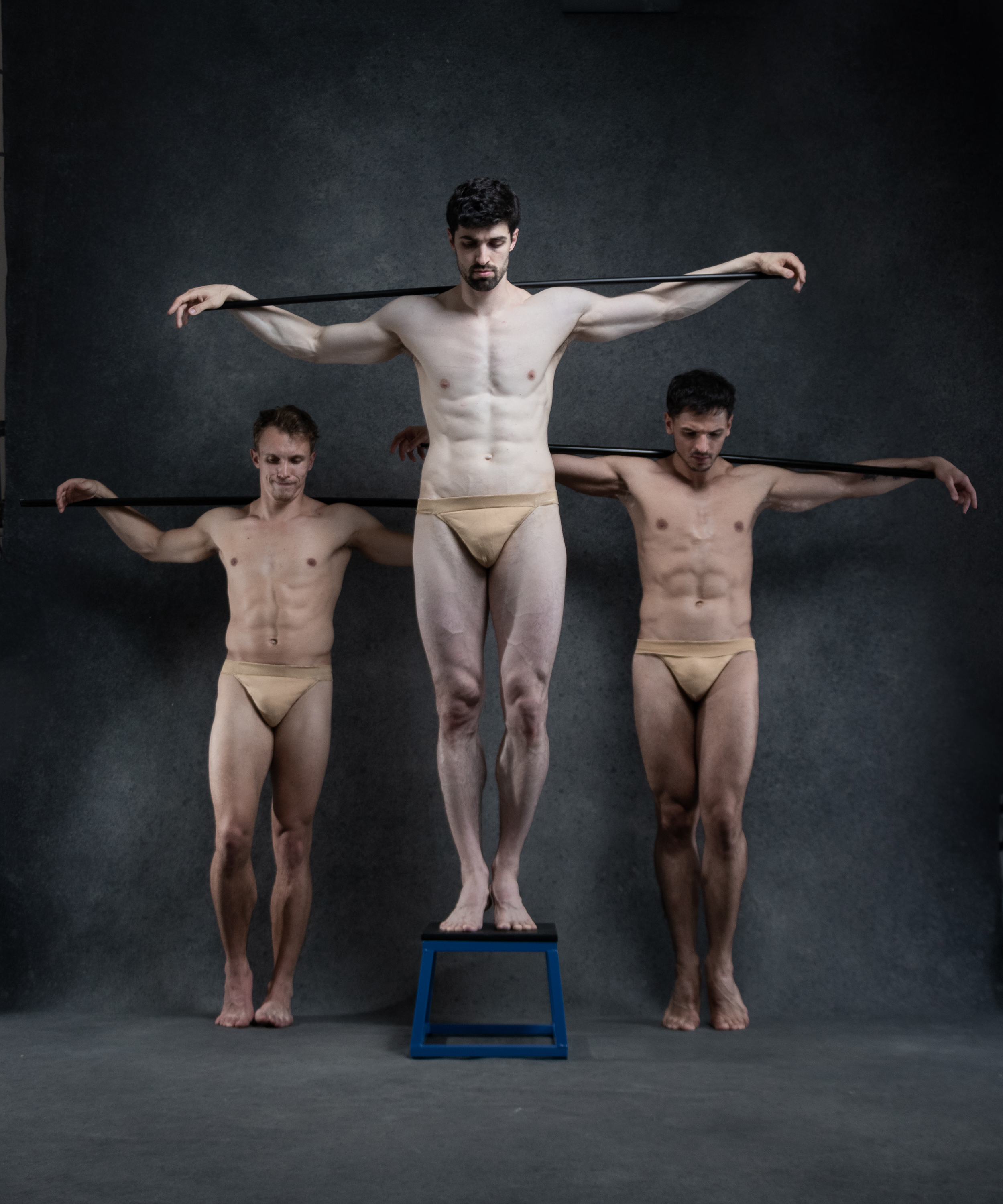 three muscular men in underwear pose in a dramatic tableau against a dark studio background.  the man in the center stands on a blue stool with his arms outstretched holding a black pole. the men on either side of him stand with their arms outstretched holding the pole as well, creating a symmetrical and striking composition. the lighting highlights their physiques, adding to the artistic and powerful nature of the image.